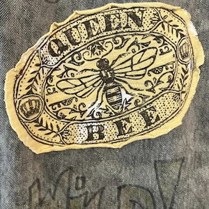 Pode incluir: Um emblema de tecido bege com bordas desfiadas apresentando uma ilustra&ccedil;&atilde;o em tinta preta de uma abelha centralizada dentro de uma moldura oval. O texto QUEEN BEE est&aacute; impresso em letras mai&uacute;sculas pretas ao redor da abelha, com coroas.