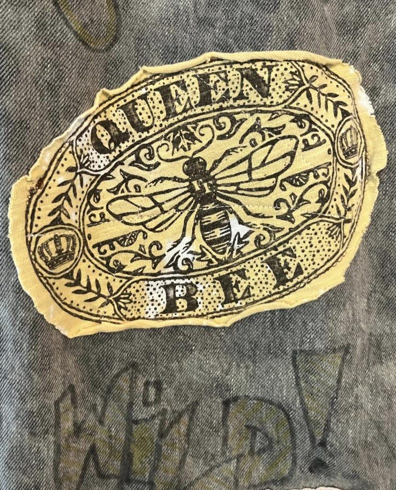 Pode incluir: Um emblema de tecido bege com bordas desfiadas apresentando uma ilustra&ccedil;&atilde;o em tinta preta de uma abelha centralizada dentro de uma moldura oval. O texto QUEEN BEE est&aacute; impresso em letras mai&uacute;sculas pretas ao redor da abelha, com coroas.