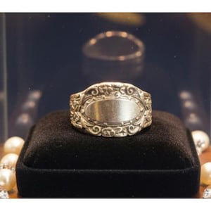Op de afbeelding: Een zilverkleurige ring met een ovaalvormige centrale plaat met een glad, gepolijst oppervlak. De band is versierd met ingewikkeld, verhoogd krulwerk en bloemachtige patronen die zich over de gehele buitenomtrek van de ring uitstrekken.