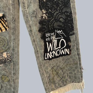 Pode incluir: Cal&ccedil;as de ganga cinza claro com lavagem &aacute;cida, pain&eacute;is de renda preta, remendos com estampas de animais e desenhos feitos &agrave; m&atilde;o. Um remendo de tecido preto exibe o texto branco "take me into the WILD UNKNOWN" com um gr&aacute;fico de montanha. Bainha desfiada.