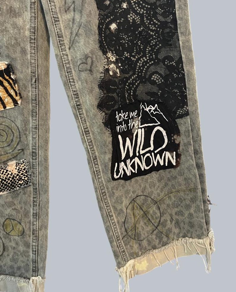 Pode incluir: Cal&ccedil;as de ganga cinza claro com lavagem &aacute;cida, pain&eacute;is de renda preta, remendos com estampas de animais e desenhos feitos &agrave; m&atilde;o. Um remendo de tecido preto exibe o texto branco "take me into the WILD UNKNOWN" com um gr&aacute;fico de montanha. Bainha desfiada.