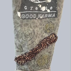 Pode incluir: Um par de cal&ccedil;as de ganga cinzentas com lavagem &aacute;cida, com um grande remendo retangular com arte floral branca e o texto "create GOOD KARMA". As cal&ccedil;as incluem uma tira diagonal de tecido com padr&atilde;o de leopardo e s&iacute;mbolos desenhados.