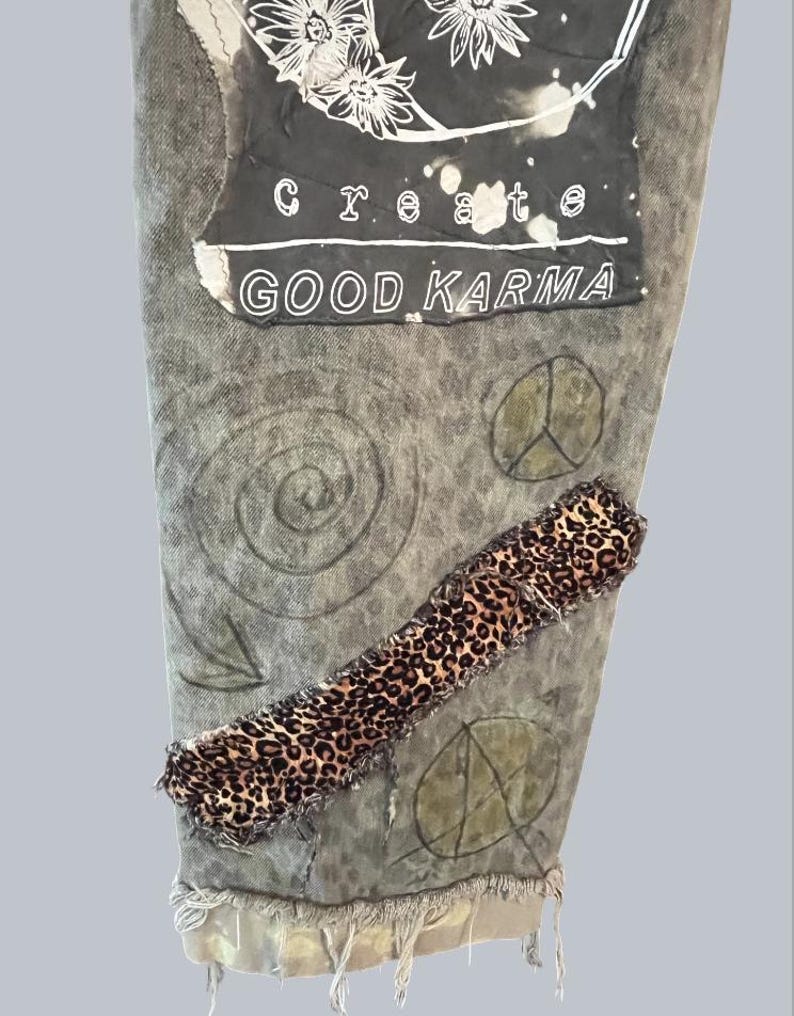 Pode incluir: Um par de cal&ccedil;as de ganga cinzentas com lavagem &aacute;cida, com um grande remendo retangular com arte floral branca e o texto "create GOOD KARMA". As cal&ccedil;as incluem uma tira diagonal de tecido com padr&atilde;o de leopardo e s&iacute;mbolos desenhados.