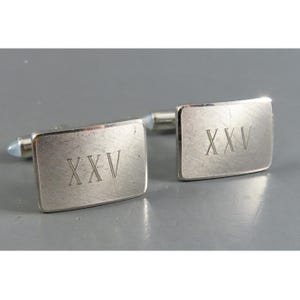 May include: A pair of rectangular silver-toned metal cufflinks with a brushed finish. Each cufflink features the Roman numeral XXV engraved in the center. The design includes a standard swivel toggle mechanism on the back for secure fastening.