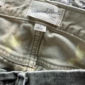 Pode incluir: Uma cal&ccedil;a jeans de cor clara com um padr&atilde;o tie-dye sutil com detalhes em amarelo. O c&oacute;s interno exibe uma etiqueta de tecido branco com o texto Universal Thread Goods Co. e uma etiqueta de tamanho 2, fabricada em Bangladesh.
