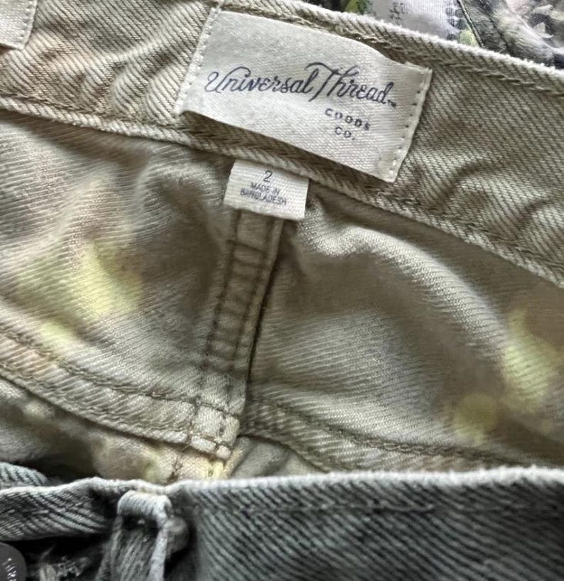 Pode incluir: Uma cal&ccedil;a jeans de cor clara com um padr&atilde;o tie-dye sutil com detalhes em amarelo. O c&oacute;s interno exibe uma etiqueta de tecido branco com o texto Universal Thread Goods Co. e uma etiqueta de tamanho 2, fabricada em Bangladesh.