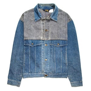 May include: A two-tone denim jacket featuring a grey upper chest panel and a blue lower section. The jacket includes a pointed collar, long sleeves, a front button closure with metallic buttons, and two chest pockets with buttoned flaps. A Wrangler brand label is visible on the inner neck.