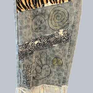 Pode incluir: Cal&ccedil;as de ganga com lavagem &aacute;cida, apresentando um remendo com estampado de tigre no topo, uma faixa horizontal com padr&atilde;o de pele de cobra a preto e branco e ilustra&ccedil;&otilde;es a tinta preta desenhadas &agrave; m&atilde;o, incluindo um rosto sorridente, uma espiral, um sinal de paz e a palavra PEACE.