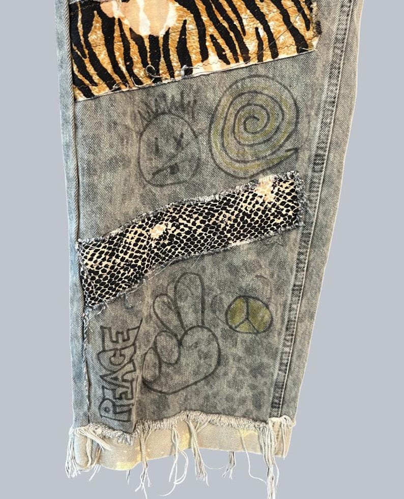 Pode incluir: Cal&ccedil;as de ganga com lavagem &aacute;cida, apresentando um remendo com estampado de tigre no topo, uma faixa horizontal com padr&atilde;o de pele de cobra a preto e branco e ilustra&ccedil;&otilde;es a tinta preta desenhadas &agrave; m&atilde;o, incluindo um rosto sorridente, uma espiral, um sinal de paz e a palavra PEACE.