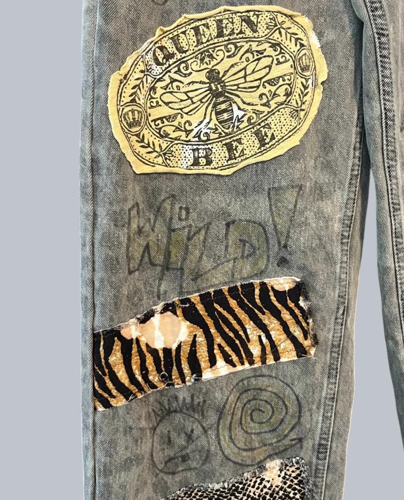 Pode incluir: Cal&ccedil;as de ganga cinzento-claro com lavagem &aacute;cida, apresentando um grande emblema oval com uma ilustra&ccedil;&atilde;o de uma abelha e o texto QUEEN BEE. Abaixo, um emblema retangular com padr&atilde;o de riscas de tigre preto e laranja, junto ao texto WILD!