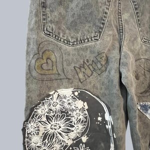 Pode incluir: Cal&ccedil;as de ganga cinzentas com lavagem &aacute;cida, apresentando um grande emblema circular com um estampado floral branco sobre um fundo preto. As cal&ccedil;as incluem um contorno de cora&ccedil;&atilde;o dourado desenhado &agrave; m&atilde;o e a palavra WILD em letra estilizada.