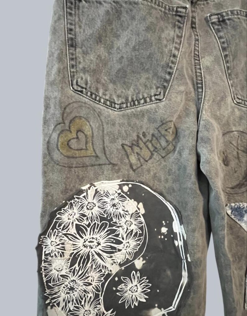 Pode incluir: Cal&ccedil;as de ganga cinzentas com lavagem &aacute;cida, apresentando um grande emblema circular com um estampado floral branco sobre um fundo preto. As cal&ccedil;as incluem um contorno de cora&ccedil;&atilde;o dourado desenhado &agrave; m&atilde;o e a palavra WILD em letra estilizada.