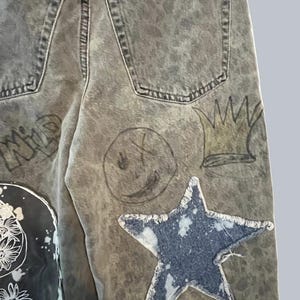 Pode incluir: Cal&ccedil;as de ganga cinzentas com lavagem &aacute;cida, com um grande emblema de estrela azul com bordas desfiadas, um smiley desenhado &agrave; m&atilde;o, uma ilustra&ccedil;&atilde;o de coroa e um emblema preto com arte floral branca. Inclui dois bolsos traseiros vis&iacute;veis.