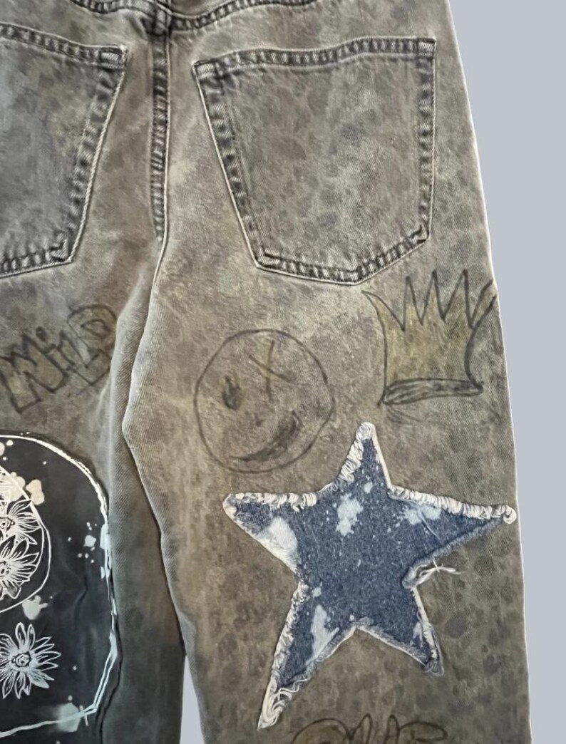 Pode incluir: Cal&ccedil;as de ganga cinzentas com lavagem &aacute;cida, com um grande emblema de estrela azul com bordas desfiadas, um smiley desenhado &agrave; m&atilde;o, uma ilustra&ccedil;&atilde;o de coroa e um emblema preto com arte floral branca. Inclui dois bolsos traseiros vis&iacute;veis.