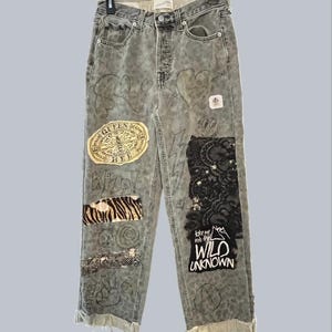 Peut inclure: Jean en denim gris à taille haute avec un imprimé léopard délavé, divers écussons dont un emblème Queen Bee et de la dentelle noire, ainsi que des motifs dessinés à la main. Le jean possède une braguette à boutons, des ourlets effilochés et le texte Take me into the WILD UNKNOWN.