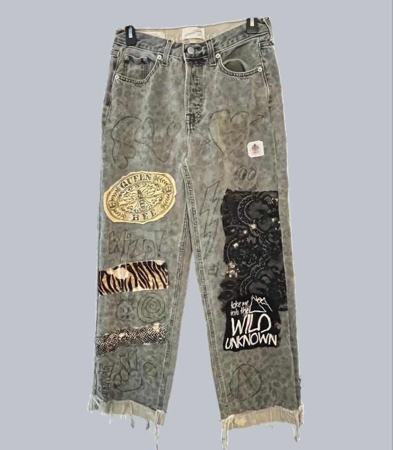 Pode incluir: Cal&ccedil;as de ganga cinzentas de cintura subida com estampado de leopardo desbotado, v&aacute;rios emblemas incluindo um emblema Queen Bee e renda preta, e desenhos feitos &agrave; m&atilde;o. As cal&ccedil;as t&ecirc;m fecho de bot&otilde;es, bainhas desfiadas e o texto Take me into the WILD UNKNOWN.