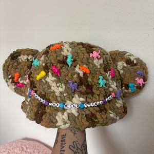 May include: A camouflage-patterned crochet bucket hat with two rounded ears on the sides. The hat is decorated with small, colorful plastic dinosaur figurines and a beaded string featuring the text WHERE THE WILD THINGS ARE in white beads.