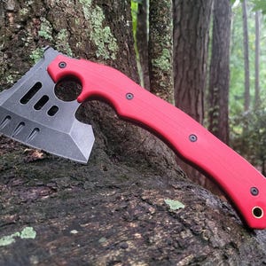 May include: A tactical hatchet featuring a matte black metal blade with three elongated cutouts and a curved, bright red handle. The handle is secured with three black screws and includes a circular finger hole and a small metal loop at the base.