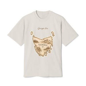 Puede incluir: Una camiseta de color beige claro con una ilustración en tinta marrón de la isla de Tasmania. El gráfico incluye una cordillera, pinos, un pequeño símbolo de corazón y el texto "Georgie Lou" arriba y "TASMANIA" centrado en el mapa.