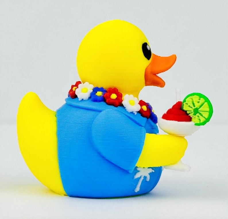 May include: A yellow rubber duck wearing a blue shirt with a white palm tree print and a colorful floral lei around its neck. The duck holds a white cocktail glass containing a red drink garnished with a bright green lime slice.