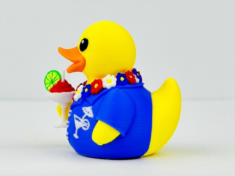 May include: A yellow rubber duck wearing a blue shirt with a white cocktail graphic print. The duck has a colorful floral lei around its neck and is holding a white cocktail glass with a red drink and a green lime slice garnish.