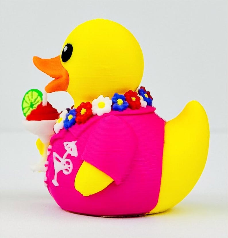 May include: A yellow rubber duck wearing a bright pink shirt with a white cocktail print, a colorful floral lei around its neck, and holding a small white cocktail glass with a red drink and a green lime slice garnish in its orange beak.