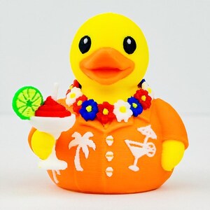 May include: A yellow rubber duck wearing an orange shirt with white palm tree and cocktail graphics. The duck has an orange beak, black eyes, and a colorful floral lei around its neck. It holds a white cocktail glass with a lime slice garnish.