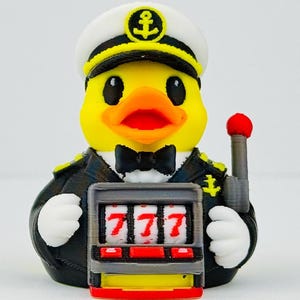 Puede incluir: Un pato de goma amarillo vestido de capit&aacute;n de barco con uniforme negro y gorra blanca con un emblema de ancla. El pato lleva una pajarita negra y sostiene una m&aacute;quina tragaperras gris con tres sietes rojos en los rodillos y una palanca.