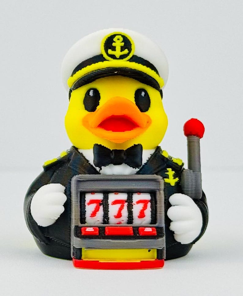 Puede incluir: Un pato de goma amarillo vestido de capit&aacute;n de barco con uniforme negro y gorra blanca con un emblema de ancla. El pato lleva una pajarita negra y sostiene una m&aacute;quina tragaperras gris con tres sietes rojos en los rodillos y una palanca.