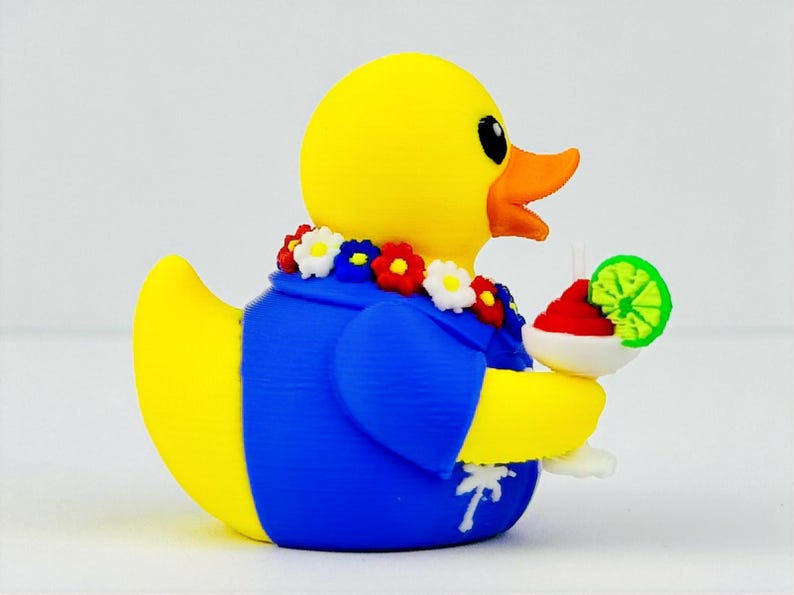 May include: A yellow rubber duck wearing a blue shirt with a white palm tree print and a colorful floral lei around its neck. The duck holds a white cocktail glass containing a red drink garnished with a bright green lime slice in its wing.