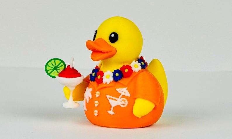 May include: A yellow rubber duck wearing an orange short-sleeved shirt with white palm tree and cocktail graphics. The duck has a floral lei around its neck and holds a white cocktail glass with a red drink, a white straw, and a green lime slice.