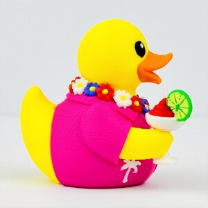 May include: A yellow rubber duck wearing a bright pink shirt with a white palm tree print and a colorful floral lei around its neck. The duck holds a white cocktail glass containing a red drink garnished with a green lime slice.