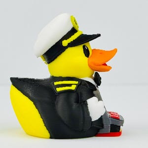 Puede incluir: Una figura de pato de goma amarillo vestido de capit&aacute;n de barco, con una gorra blanca y negra con adornos amarillos, una chaqueta negra con charreteras amarillas, una pajarita negra y sosteniendo un dispositivo gris con botones rojos.