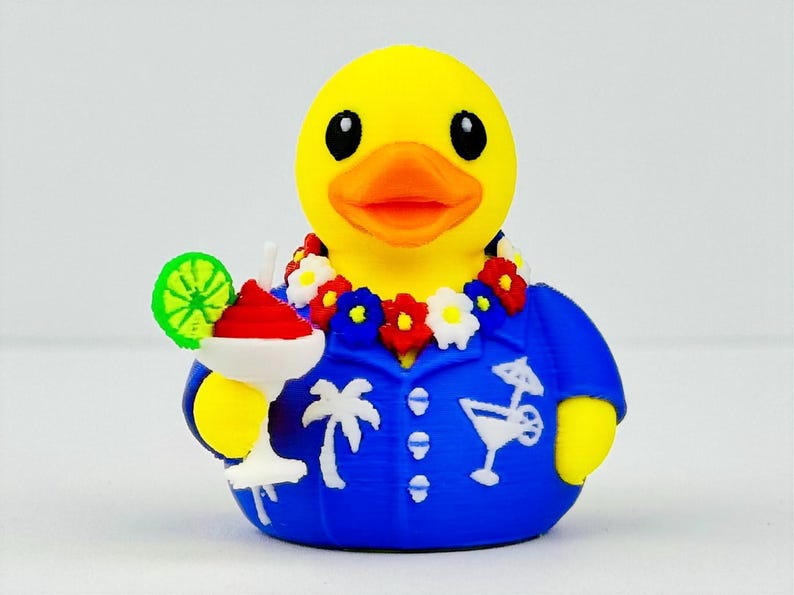 May include: A yellow rubber duck wearing a blue button-down shirt with white palm tree and cocktail glass patterns. The duck has a floral lei around its neck and is holding a white cocktail glass with a red drink and a green lime slice garnish.