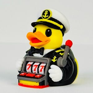 Puede incluir: Un pato de goma amarillo vestido de capit&aacute;n de barco con uniforme negro y gorra blanca con un emblema de ancla. El pato sostiene una m&aacute;quina tragaperras gris con tres sietes rojos en los rodillos y una palanca con punta roja al costado.