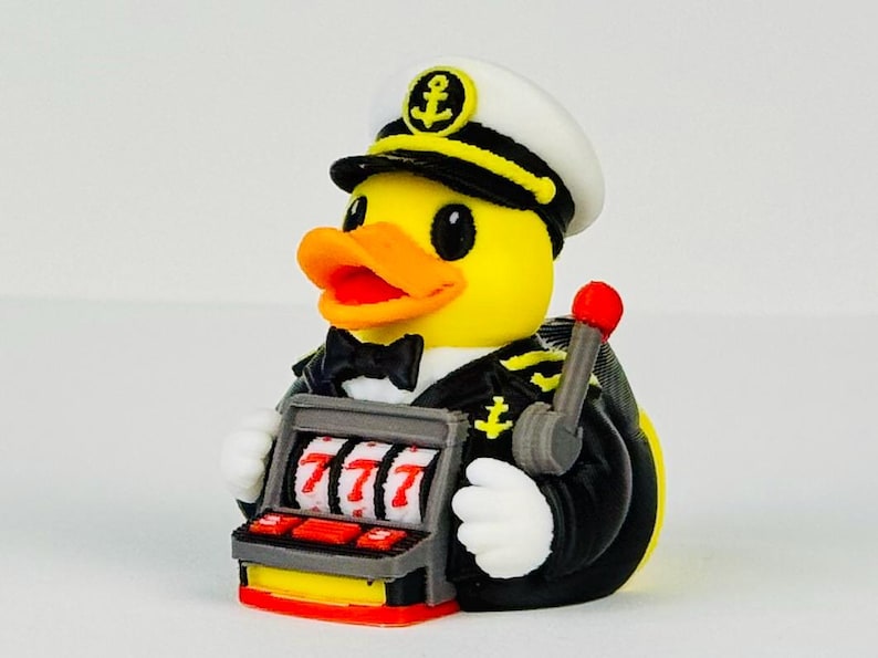 Puede incluir: Un pato de goma amarillo vestido de capit&aacute;n de barco con uniforme negro y gorra blanca con un emblema de ancla. El pato sostiene una m&aacute;quina tragaperras gris con tres sietes rojos en los rodillos y una palanca con punta roja al costado.