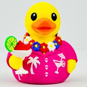 May include: A yellow rubber duck wearing a bright pink shirt with white palm tree and cocktail graphics. The duck has an orange beak, black eyes, a colorful floral lei around its neck, and holds a white cocktail glass with a red drink and lime.