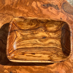 Puede incluir: Un cuenco rectangular de madera de olivo, que presenta un patrón de veta natural prominente y complejo con tonos arremolinados de marrón oscuro y amarillo dorado en toda la superficie lisa y pulida del interior y el exterior.