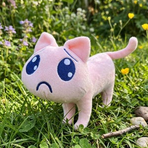 Puede incluir: Un juguete de peluche de gato rosa claro con una cabeza grande, orejas puntiagudas y una cola larga. El gato presenta ojos azul oscuro bordados con reflejos blancos y una expresión de boca triste hacia abajo. El peluche tiene una textura suave.