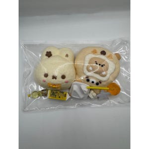 May include: A clear plastic bag containing two beige, rounded, character-shaped charms with brown facial features and small decorative accents. Included are miniature accessories: a yellow bottle, a yellow rectangular box, a white square, and a yellow lollipop.