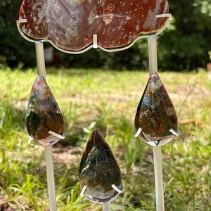 May include: A decorative metal stand shaped like a cloud with three hanging raindrops, all crafted from polished stone. The cloud is a deep reddish-brown with white speckles, while the three teardrop-shaped stones feature a mix of green, brown, and red tones.