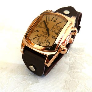 Peut inclure: Une montre-bracelet rectangulaire avec un boîtier doré et un bracelet en cuir marron. Le cadran de la montre est beige avec des chiffres et des aiguilles noirs. Le texte "WoMaGe 8088" est visible. Un accessoire élégant.