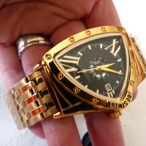 May include: A gold-toned wristwatch with a black face and gold hands. The watch has a unique triangular shape with Roman numerals and a date display. The watch band is also gold-toned. The word "BESTWIN" is printed on the watch.