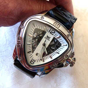 Peut inclure: Une montre-bracelet triangulaire argentée avec un cadran blanc et un bracelet en cuir noir. La montre présente un design unique avec des mécanismes internes visibles et un cadran texturé. Le bracelet est en cuir noir.