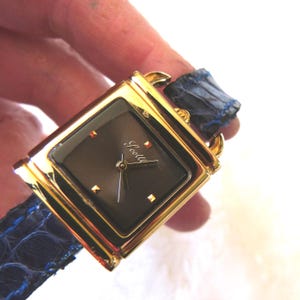 Può includere: Un orologio da polso color oro con quadrante quadrato scuro e cinturino in pelle blu. Il quadrante dell'orologio ha indicatori color oro e il nome del marchio "Seakill" in corsivo. Il cinturino è blu scuro con un motivo strutturato.