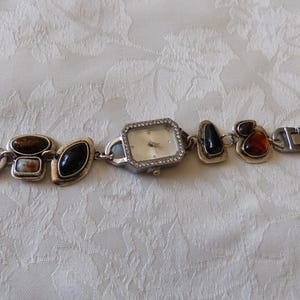 Könnte beinhalten: Eine silberne Uhr mit weißem Zifferblatt und silbernem Armband. Das Armband ist mit verschiedenen farbigen Steinen verziert.