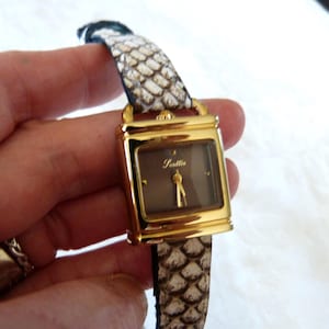 Può includere: Un orologio dorato con quadrante rettangolare e cinturino a fantasia serpente. Il quadrante è marrone tenue con lancette dorate e la scritta "Seattle". Il cinturino è un mix di bianco, beige e nero.