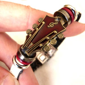 May include: A music-themed bracelet featuring a guitar head pendant in burgundy and gold tones. The bracelet has a black leather band with silver and red accents. The guitar head has a treble clef symbol.