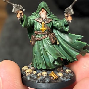 Może przedstawiać: Miniaturowa figurka brodatej postaci w poszarpanej zielonej szacie z kapturem, trzymającej dwa srebrne pistolety. Postać ma brązowy pas z sakiewkami i złoty wisiorek w kształcie krzyża. Podstawa jest ozdobiona wieloma małymi czaszkami i gruzem.