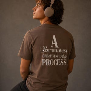 May include: A brown short-sleeved t-shirt featuring white text on the back that reads A BEAUTIFUL MESSY CREATIVE & HOLY PROCESS. The shirt is worn with light brown over-ear headphones and black trousers, showcasing a casual and simple design.